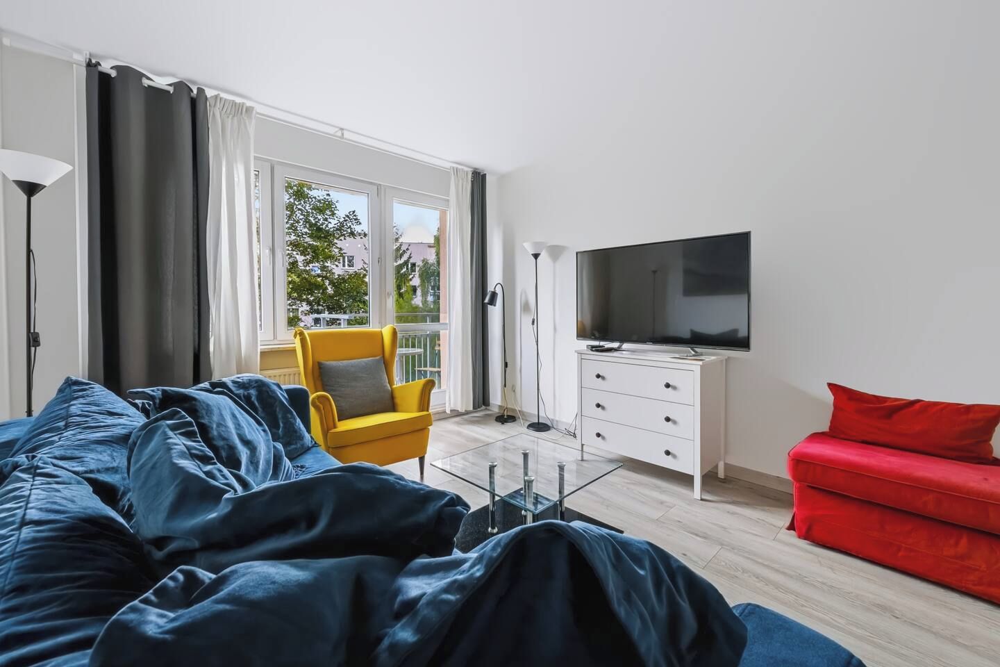 Ein modernes Wohnzimmer mit einem großen Bett, einem roten Sofa, einem gelben Sessel, einem Fernseher und einem Glastisch.
