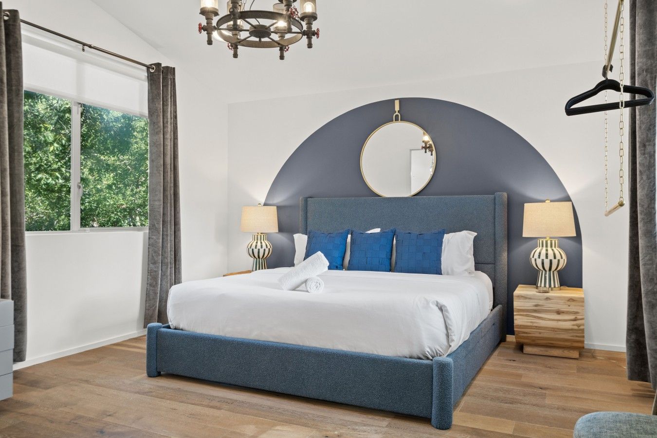 A bedroom with a large bed, two bedside tables with lamps, a round mirror above the bed, and a view of greenery outside.