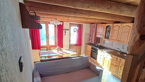 Un interior acollidor amb un sofà, sostre de fusta, zona de cuina i una finestra amb cortines vermelles.