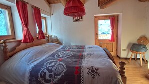 A bedroom with a wooden bed, a red hanging light fixture, and a view of the outside through a window with red curtains. - Chalet 'Lo Pailler De Nonno' with Mountain View, Shared Garden, and Wi-Fi (Maisonasse)