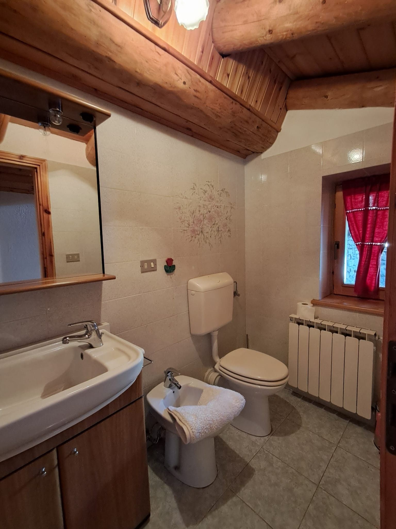 Un bagno con soffitto in legno, un lavandino, un water e una finestra con tenda rossa.