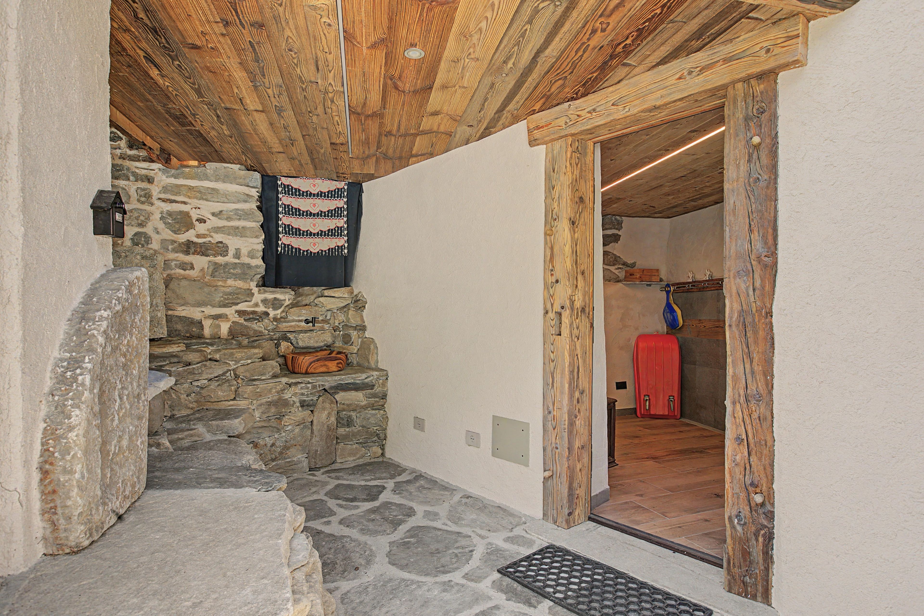 A stone staircase leading to a wooden ceiling, a stone wall with a hanging cloth, and a red suitcase in a doorway.