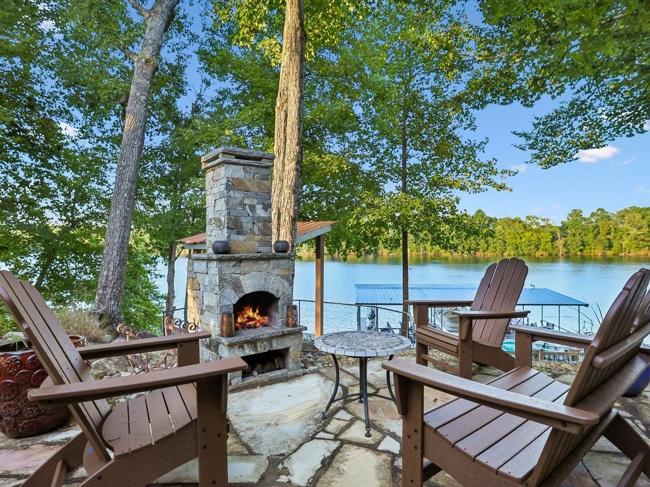 An outdoor seating area with Adirondack chairs, a stone fireplace, and a view of a lake.