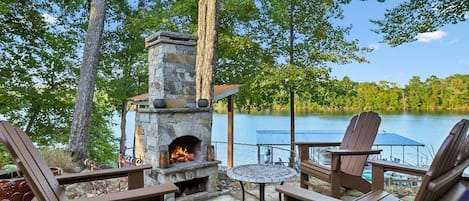 An outdoor seating area with Adirondack chairs, a stone fireplace, and a view of a lake.