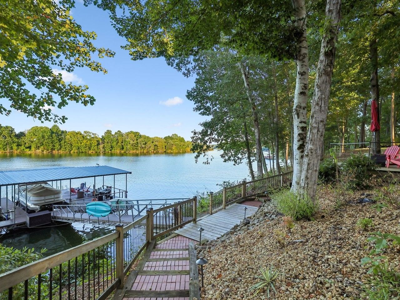 Une terrasse au bord du lac avec un espace salon couvert, un quai avec un bateau et une passerelle en bois menant à une chaise rouge.