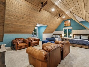 Une chambre à coucher aménagée dans un grenier, avec un plafond en pente, des poutres en bois, une grande fenêtre et un lit doté d’une tête de lit.