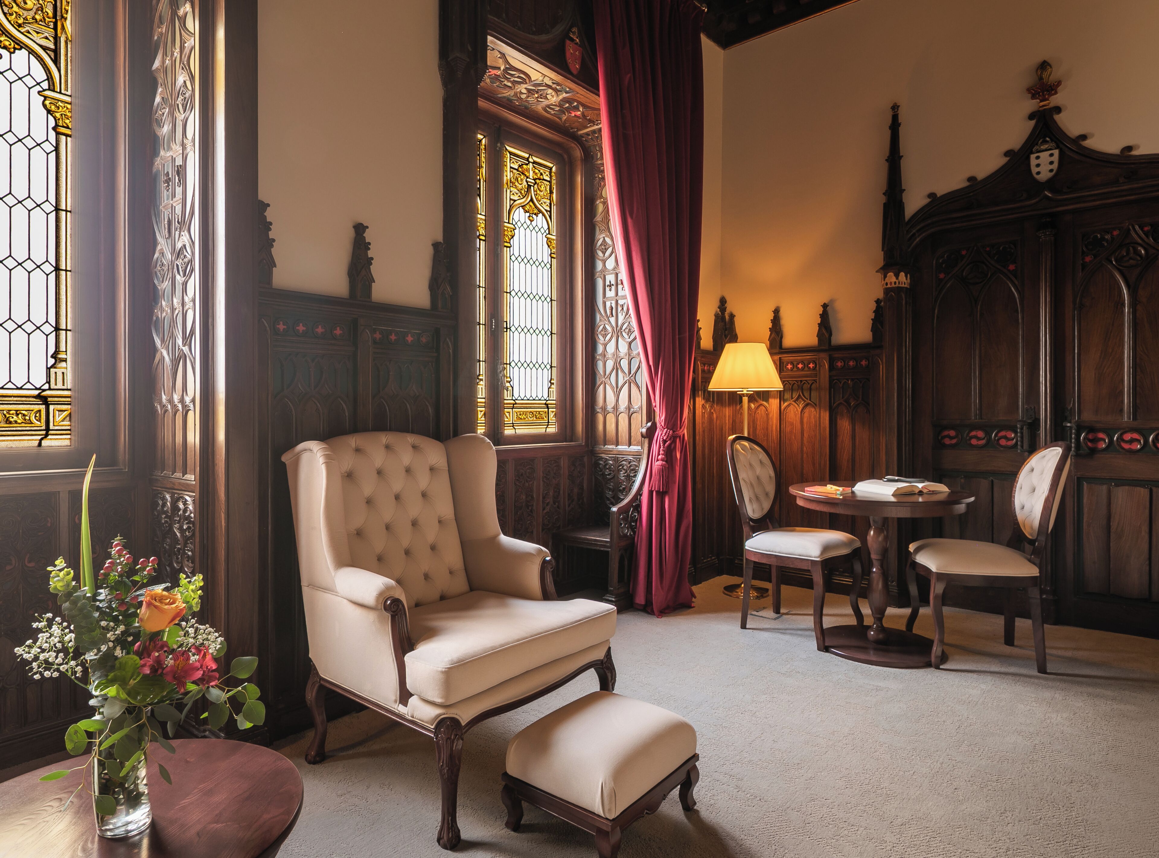 Una habitación con un sillón grande, una mesa con sillas, un florero con flores y una lámpara.