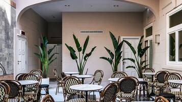 Un amplio espacio exterior con varias mesas y sillas de mimbre, rodeado de grandes ventanales y plantas en macetas.