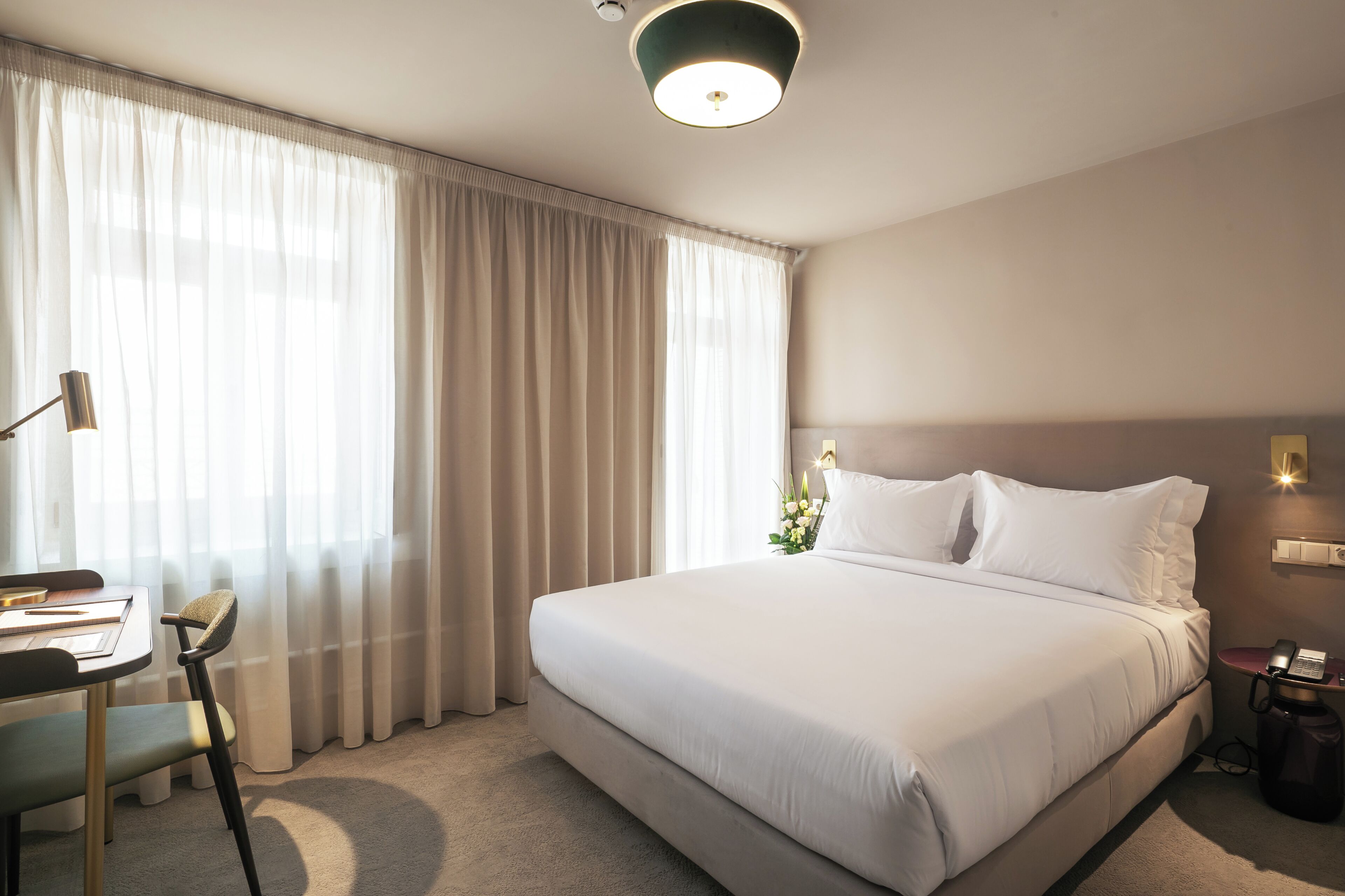 Habitación de hotel con cama, escritorio, silla, lámpara y mesita de noche con teléfono.