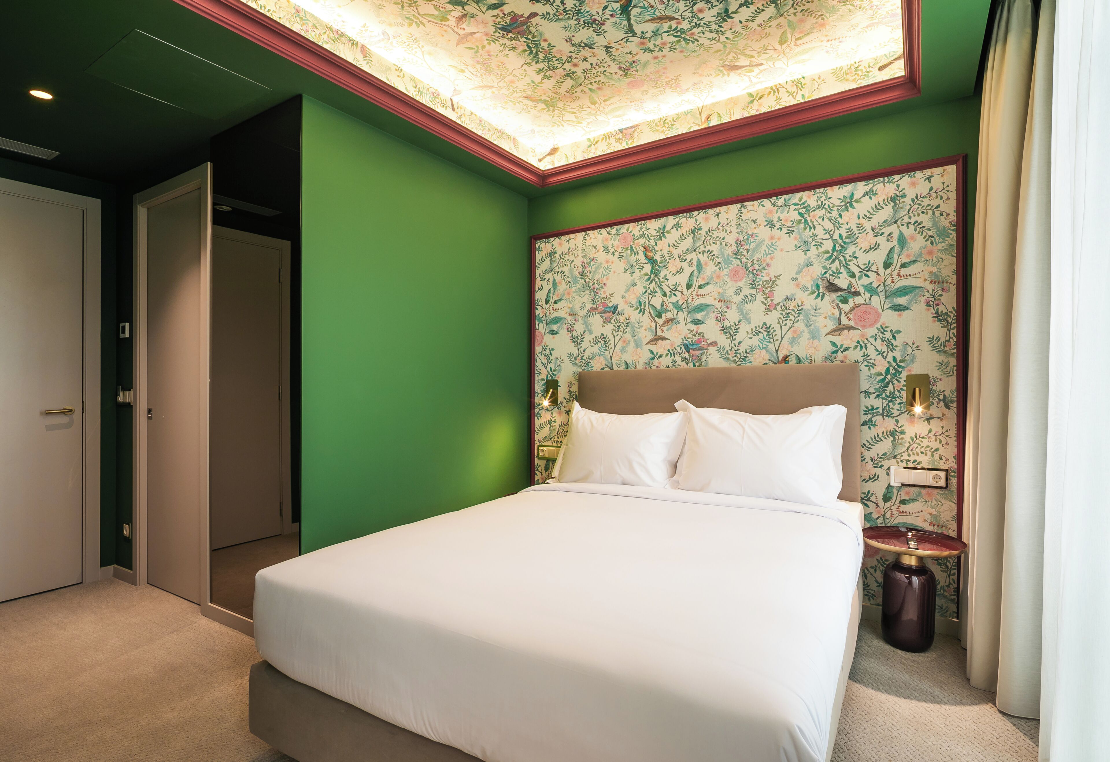 Una habitación de hotel con una cama, paredes verdes, papel tapiz con motivos florales y una lámpara en el techo.