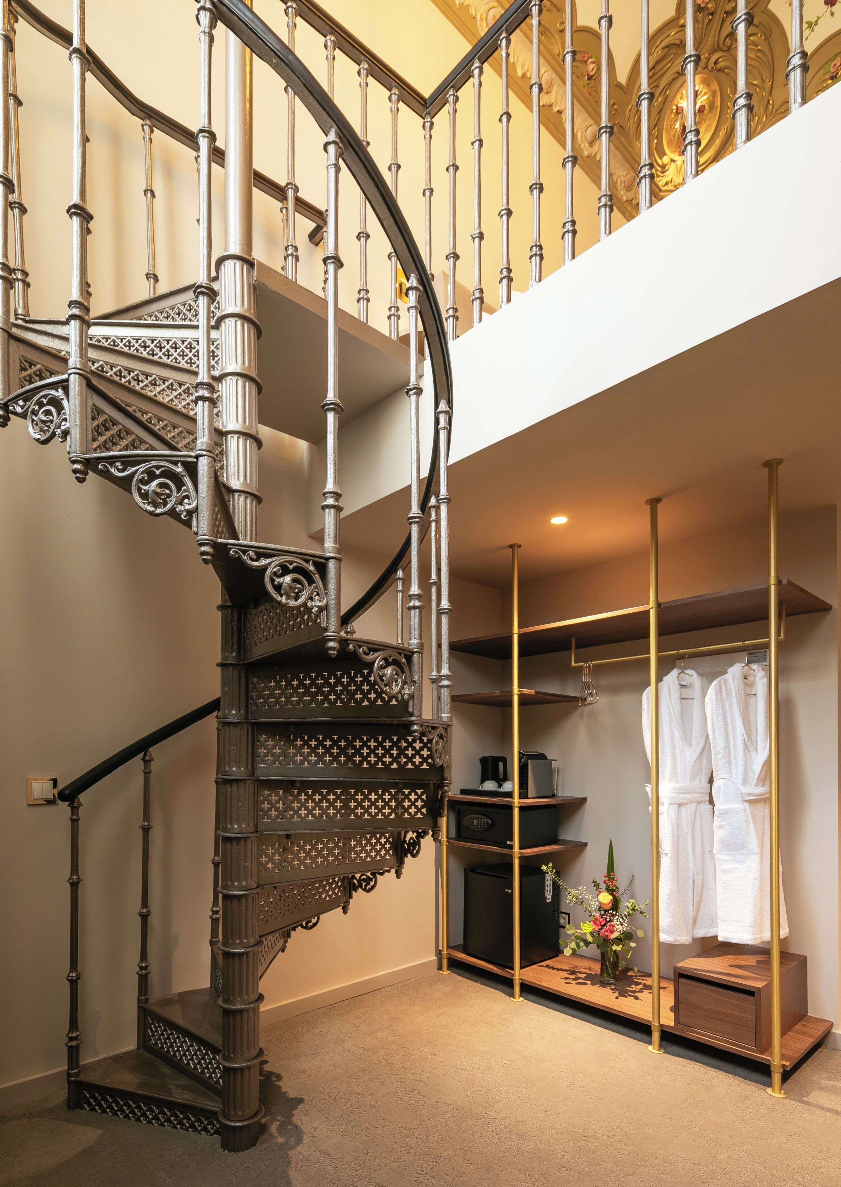 A spiral staircase with ornate metal railings and a wooden finish, leading to a second-floor area with a built-in wardrobe and a small seating area.