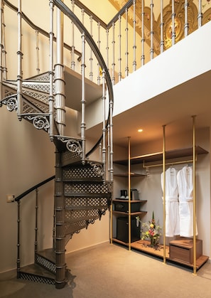 Una escalera de caracol con barandillas metálicas ornamentadas y detalles de madera, que conduce a una zona del segundo piso con un armario empotrado y un pequeño rincón de descanso.