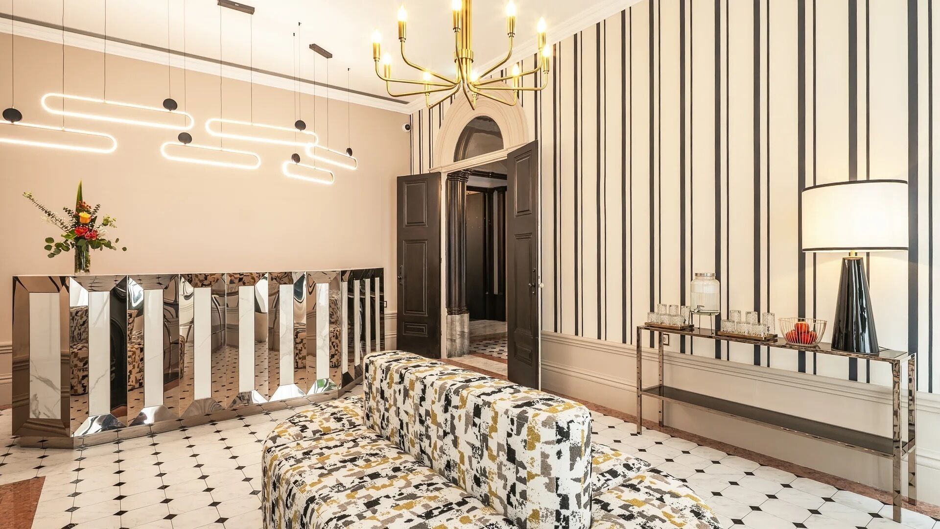 A hotel lobby with a patterned sofa, a reflective mirror wall, a chandelier, and a tiled floor.