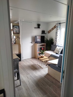 Ein kompakter Wohnbereich mit Küchenzeile, einer Sitzecke mit Sofa und Couchtisch, einem Fernseher und einem Fenster mit Vorhängen.