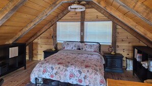 Un dormitorio tipo loft con techo inclinado, una cama con ropa de cama floral, mesitas de noche y una ventana con persianas.
