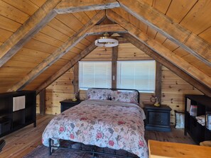 Ein Schlafzimmer im Loft-Stil mit schrägem Dach, einem Bett mit floralem Bettzeug, Nachttischen und einem Fenster mit Jalousien.