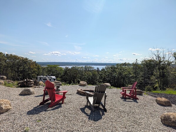A gravel area with red Adirondack chairs and a table, a white RV, and a scenic view of a lake and mountains.