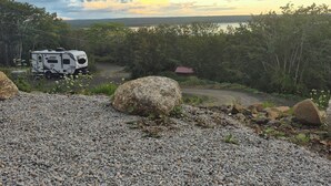 Une zone de gravier où est garavané un camping-car, le tout bordé d’arbres, avec une vue lointaine sur une étendue d’eau sous un ciel nuageux.