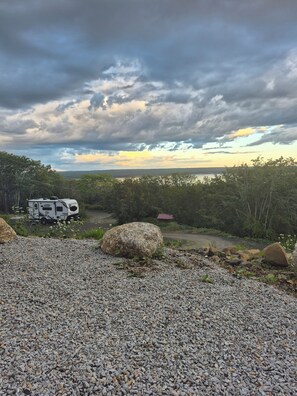 A gravel area with a camper parked on a road, surrounded by trees and a distant view of a body of water under a cloudy sky.
