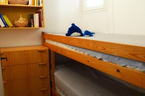 Een stapelbed met een houten hoofdbord, een kleine houten dressoir met lades, en een boekenplank met boeken en een gevlochten mand.