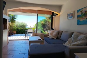 A living room with a sofa, a table, and a chair, a large window with a view of greenery and a building, and a painting on the wall. - Bright Apartment by the Ligurian sea (La croix-Valmer)