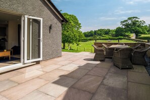 Een terras met tuinmeubilair, een stenen muur en uitzicht op een groen landschap met bomen en een hek.