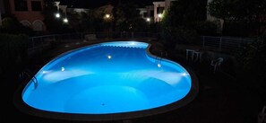 A swimming pool at night with a curved edge, surrounded by a low fence and some vegetation. - Casa Luca in the Borghi of Villasimius  (Sardegna)