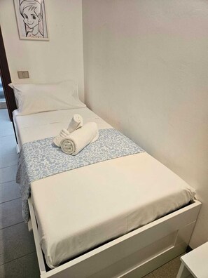 A single bed with a white mattress and a blue patterned bedspread, a rolled towel on the bed, and a wall with a framed picture. - Casa Luca in the Borghi of Villasimius  (Sardegna)