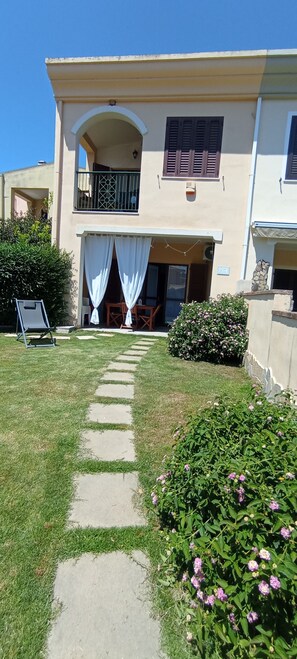 A two-story building with a covered patio, a garden with stepping stones, and a chair. - Casa Luca in the Borghi of Villasimius  (Sardegna)