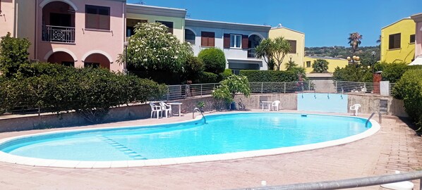 A swimming pool with a tiled edge, surrounded by a brick patio. There are white plastic chairs and a table near the pool. - Casa Luca in the Borghi of Villasimius  (Sardegna)