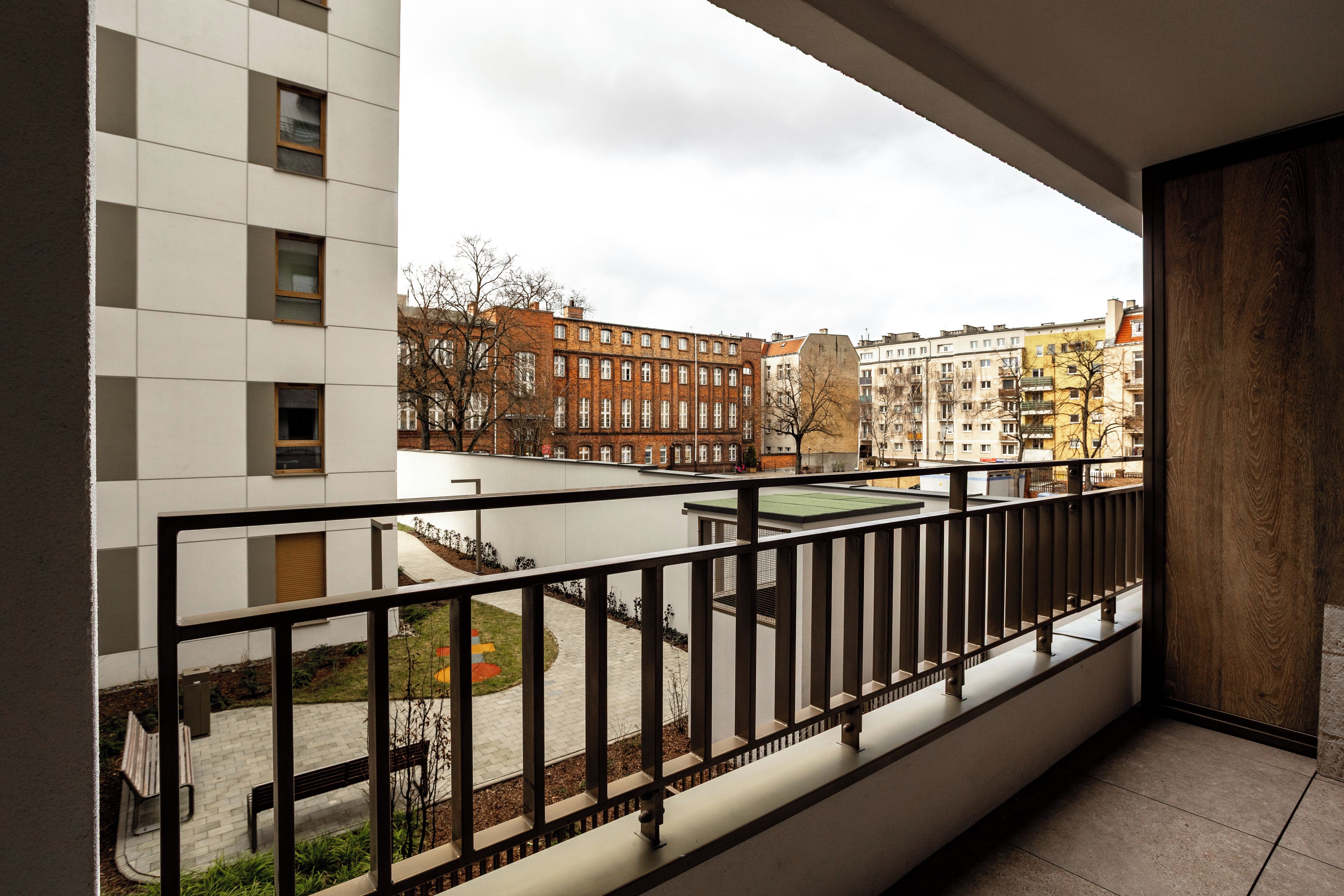 Un balcón con vista a un paisaje urbano, que incluye edificios y una zona pavimentada con bancos.