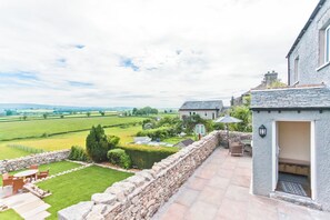 A stone wall, a patio with a table and chairs, and a view of a green field and buildings in the distance. - Hillberry Cottage (Carnforth)