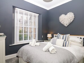 A bedroom with a bed, a heart-shaped wall decoration, a window with blinds, and a bedside table with a lamp. - 12 Courtyard Cottage at Sheplegh Court (Totnes)