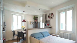 Een kleine slaapkamer met een bed, een bureau en een stoel. Er is een raam met zonwering, een radiator en een klok aan de muur.