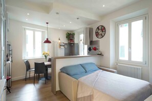 A compact bedroom with a bed, a desk, and a chair. There is a window with blinds, a radiator, and a clock on the wall. - Vrbo Property (Firenze)