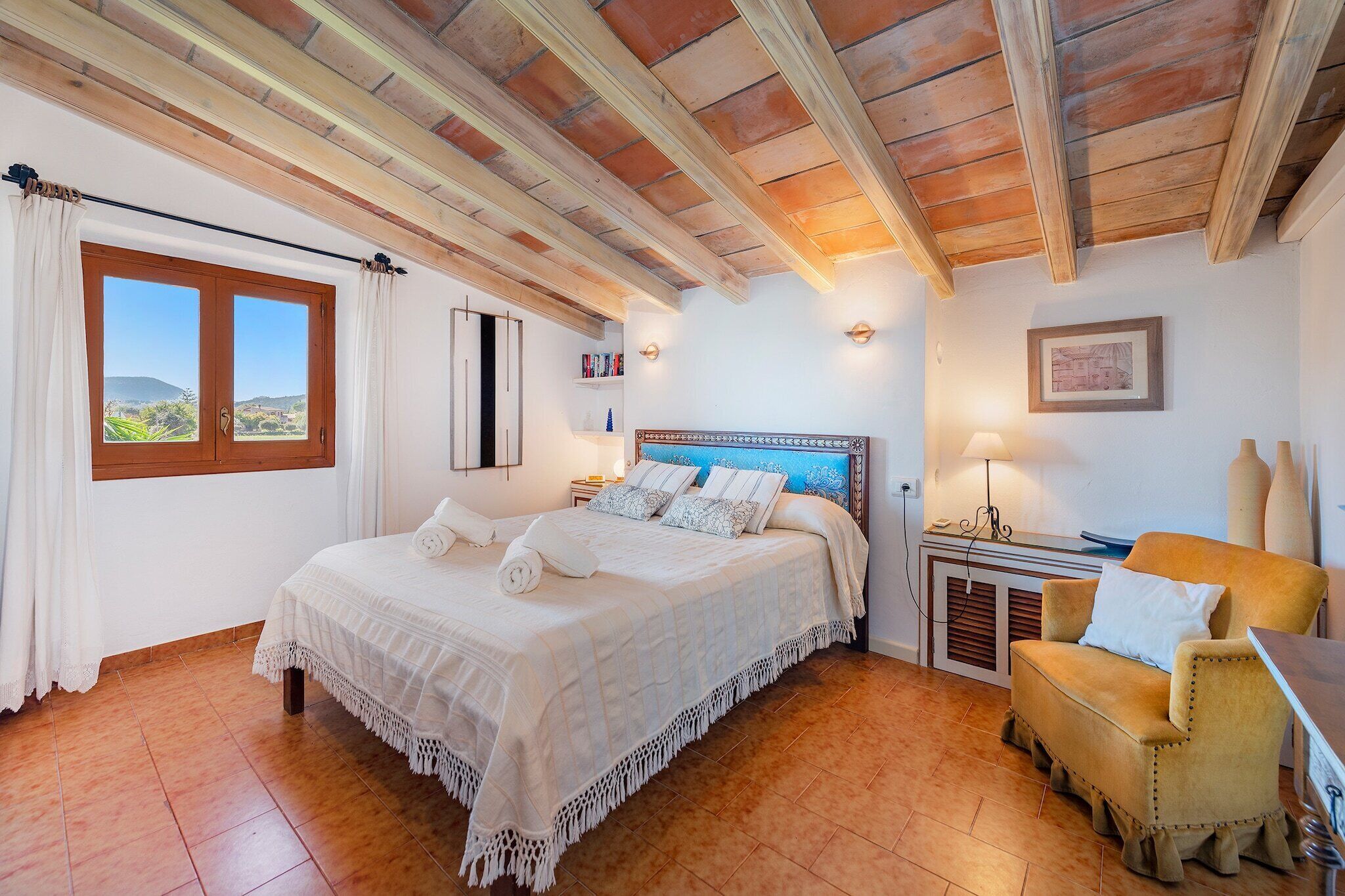 A bedroom with a large bed, a window with curtains, a bedside table, a chair, and a wall-mounted lamp.