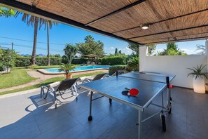 An outdoor table tennis table with a net, chairs, and a ping pong paddle, set against a backdrop of a swimming pool and greenery. - Pollensa Country Villa for 6 (Pollensa / Pollença)