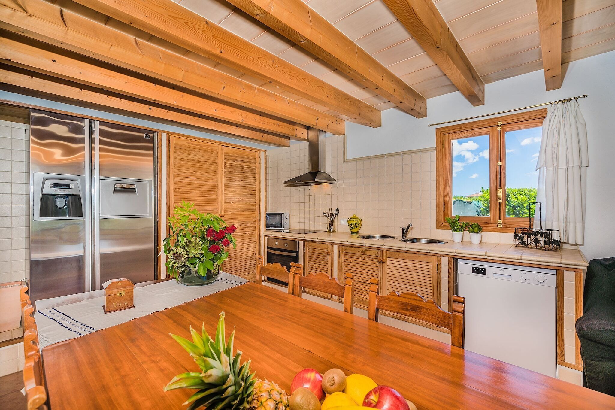A modern kitchen with wooden beams, a dining table with fruits, and a view of greenery outside.