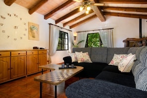 Ein gemütliches Wohnzimmer mit Holzboden, einer Couch mit floralen Kissen, einem Schachtisch und einem Holzschrank.