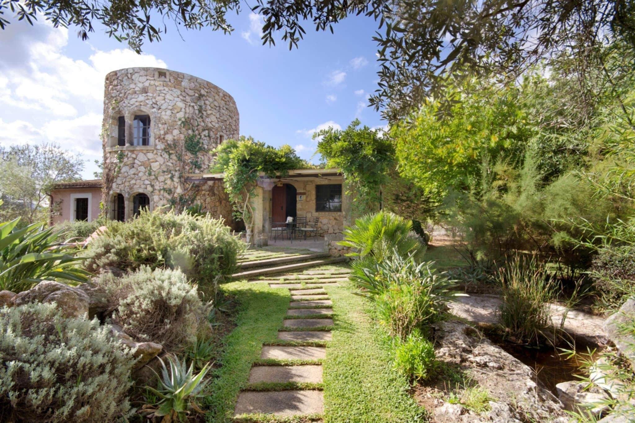 A stone building with a circular tower, surrounded by a garden with various plants and a stone pathway leading to the entrance.