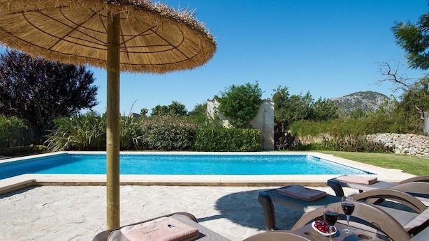 Piscina com espreguiçadeiras e um guarda-sol, cercada por vegetação e um muro de pedra.