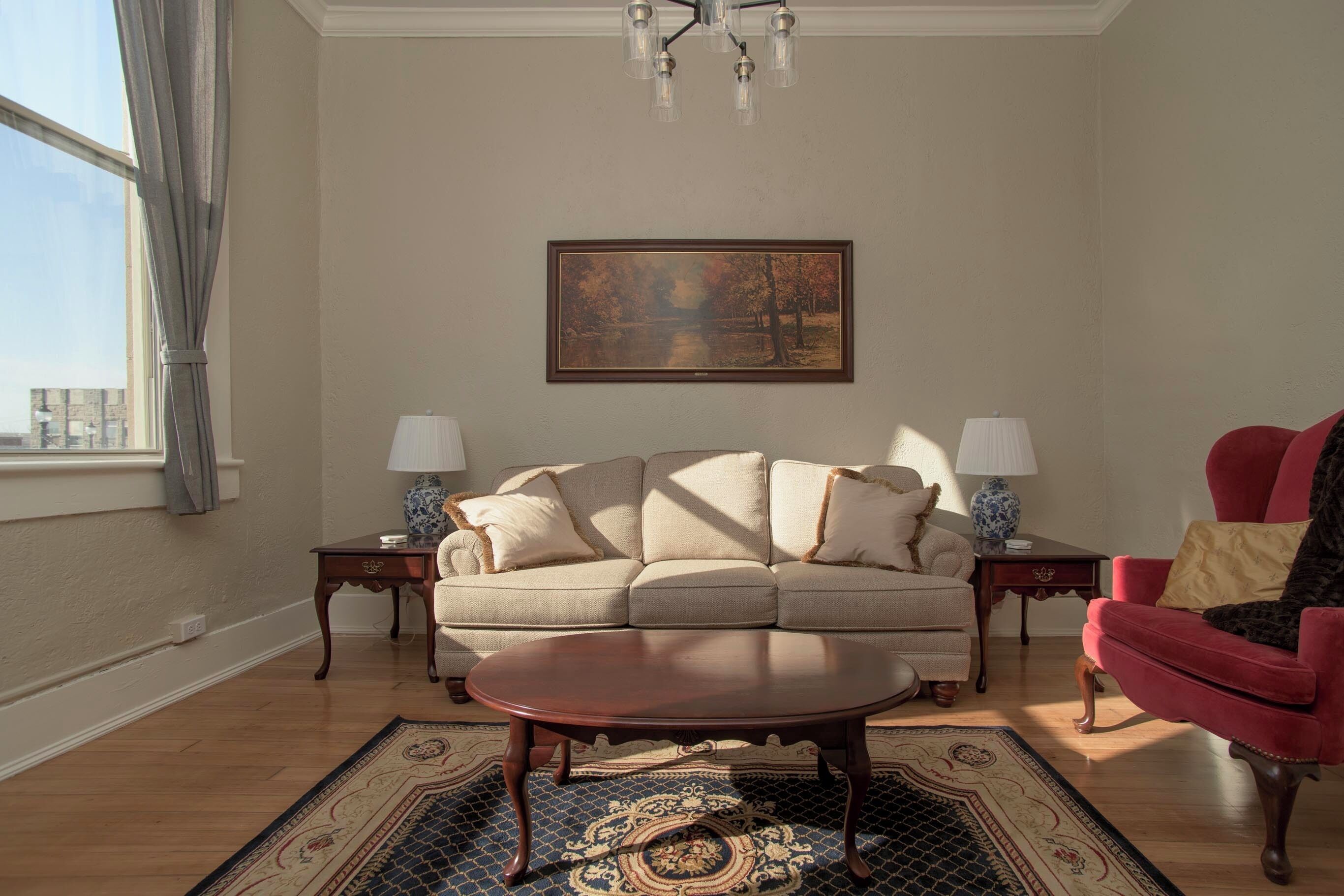 A living room with a sofa, two side tables with lamps, a red armchair, a coffee table, and a painting on the wall.
