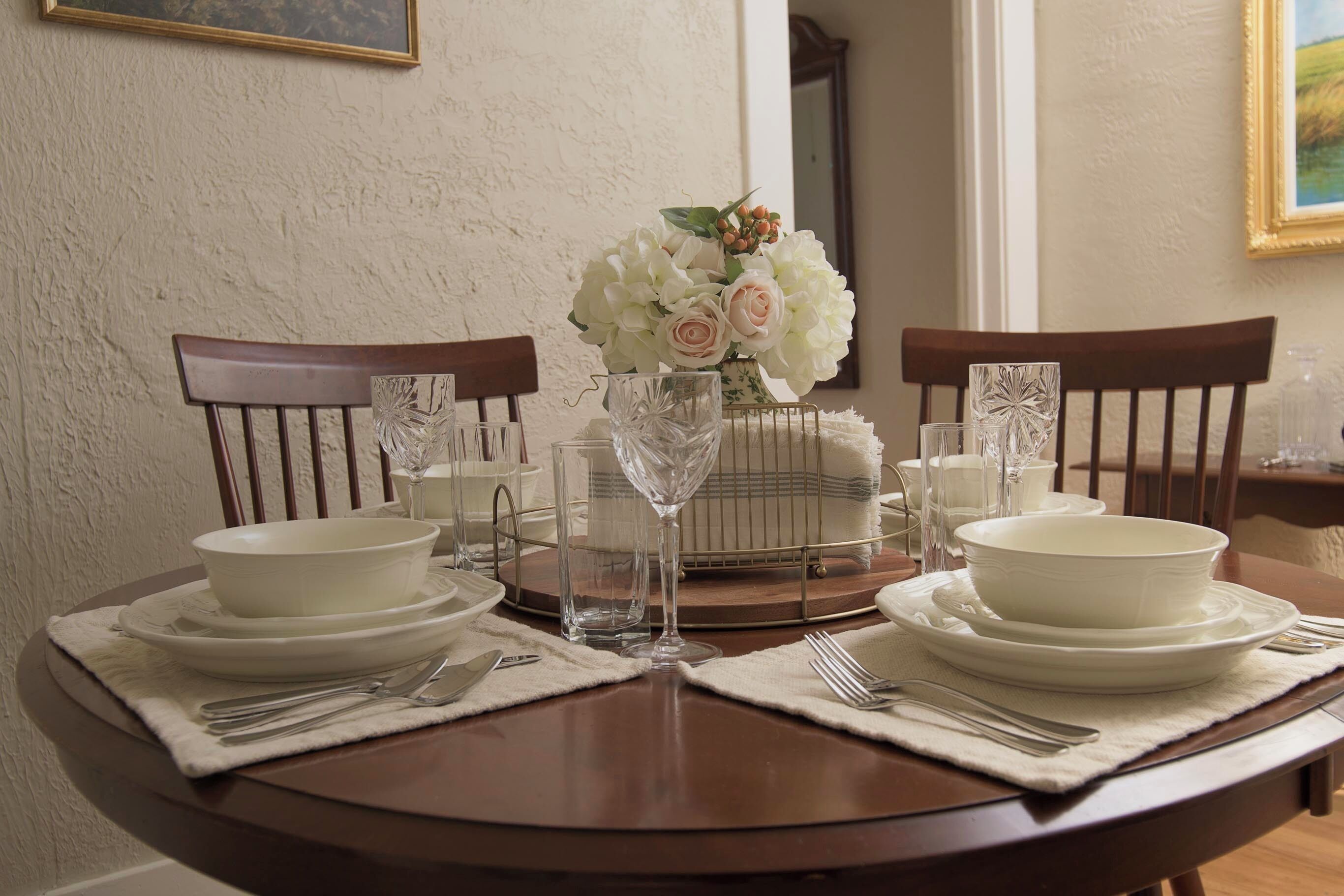 A dining table set with white plates, bowls, cutlery, and a vase of flowers.