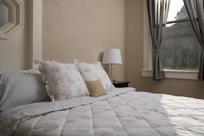 A neatly made bed with a quilted duvet and striped pillows, a bedside lamp, and a view of a building through a window with curtains.
