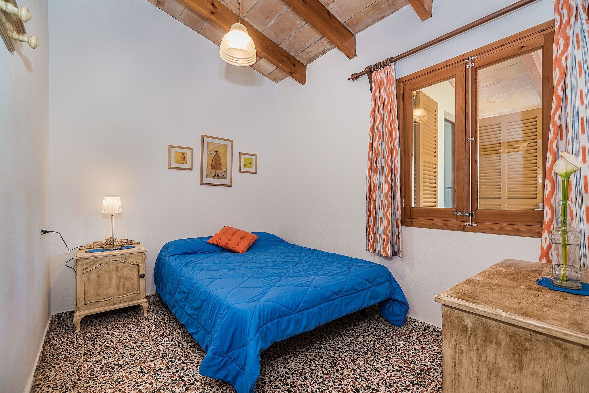 Un dormitorio con cama, mesita de noche, lámpara, ventana con cortinas y una obra de arte en la pared.