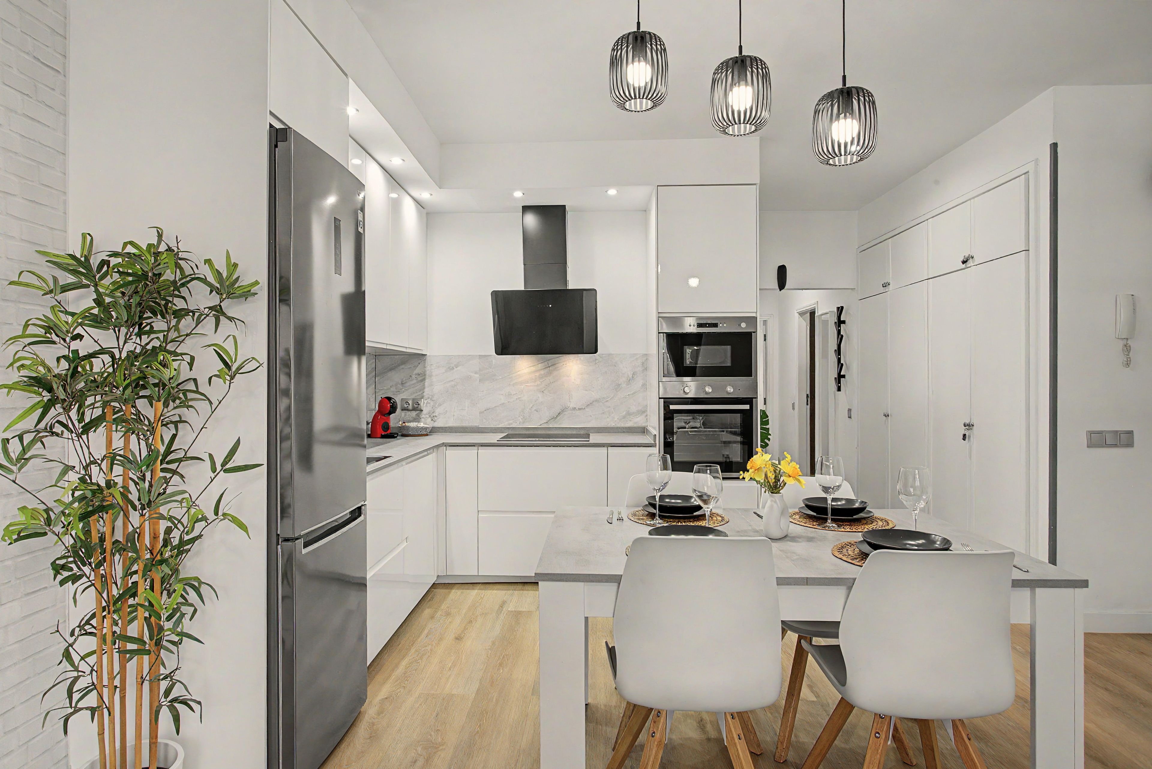 Una cocina moderna con armarios blancos, un refrigerador de acero inoxidable, un horno empotrado y un comedor con una mesa dispuesta para dos personas.