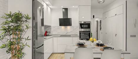Una cocina moderna con armarios blancos, un refrigerador de acero inoxidable, un horno empotrado y un comedor con una mesa dispuesta para dos personas.