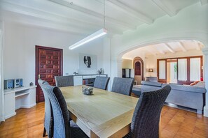 Ein Essbereich mit Holztisch und Korbstühlen, im Hintergrund eine Küche und eine gemütliche Sitzecke mit Sofa und Vorhängen.