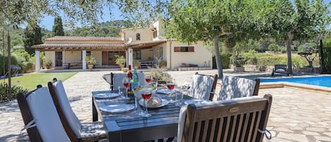 Un área de comedor al aire libre con piscina, piso de piedra y un edificio al fondo.