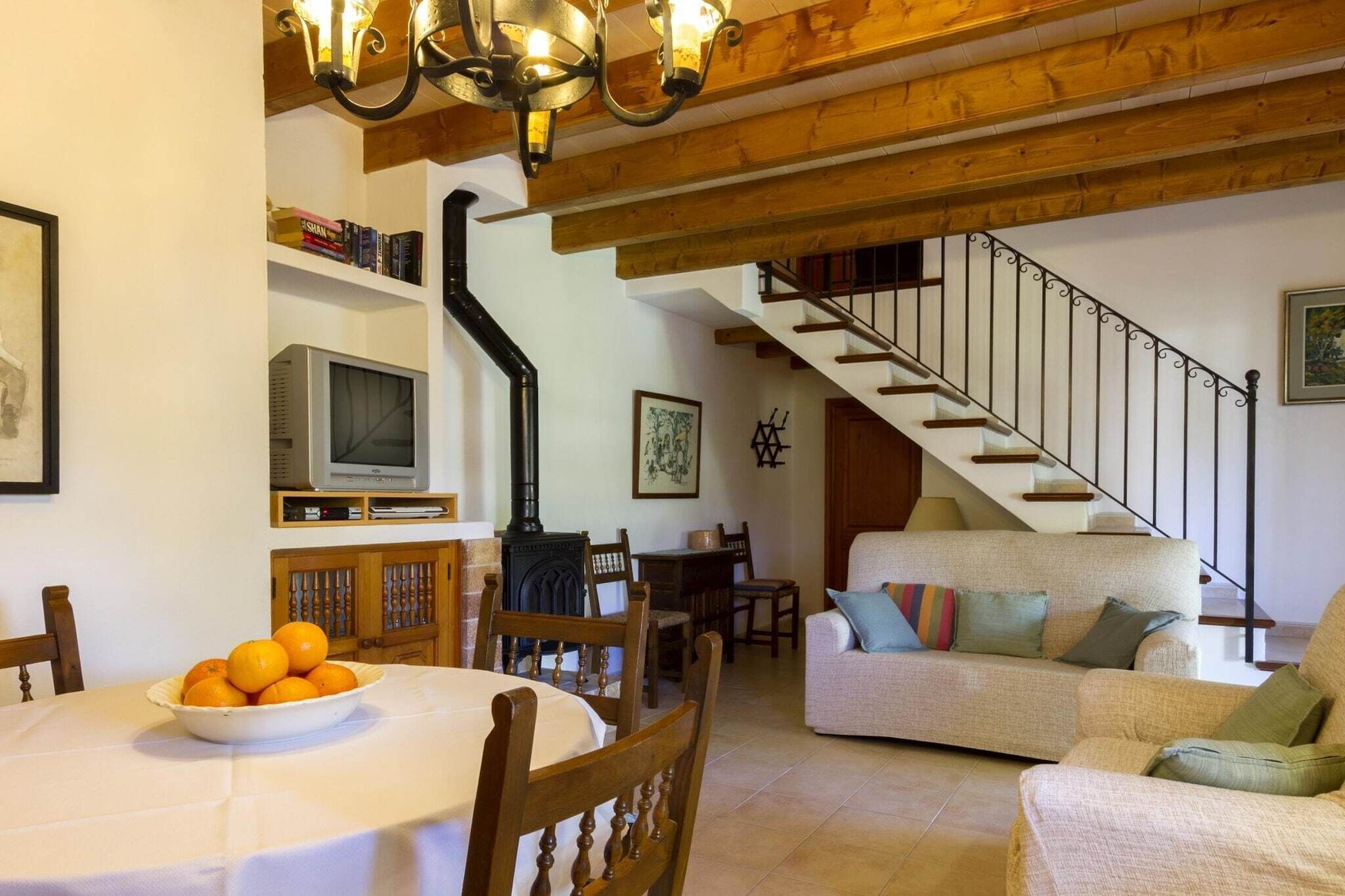 A cozy living room with a staircase, a dining area with a TV, and a bowl of oranges on the table.